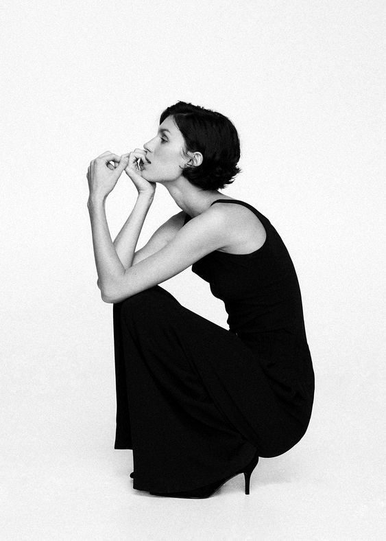 A person in a black dress crouching in profile, hands at their mouth, thinking.