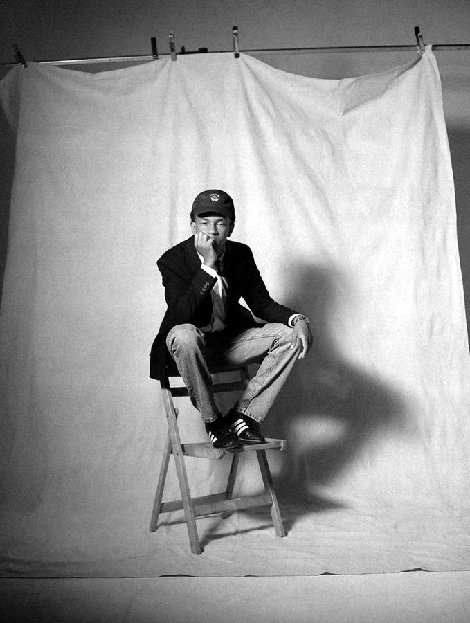 A young musician in a cap and blazer perched on a wooden chair in front of a hanging backdrop.