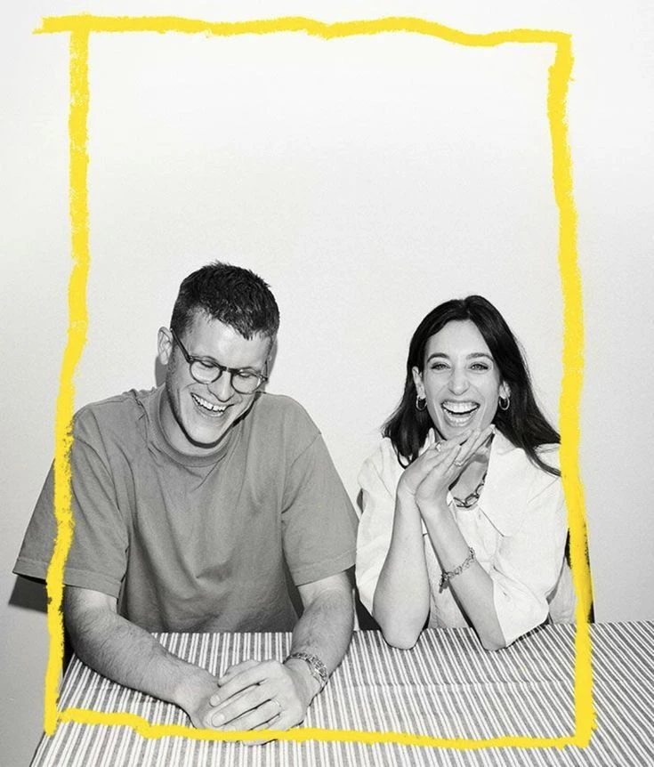 Two people laughing at a striped table inside a hand-drawn yellow frame.