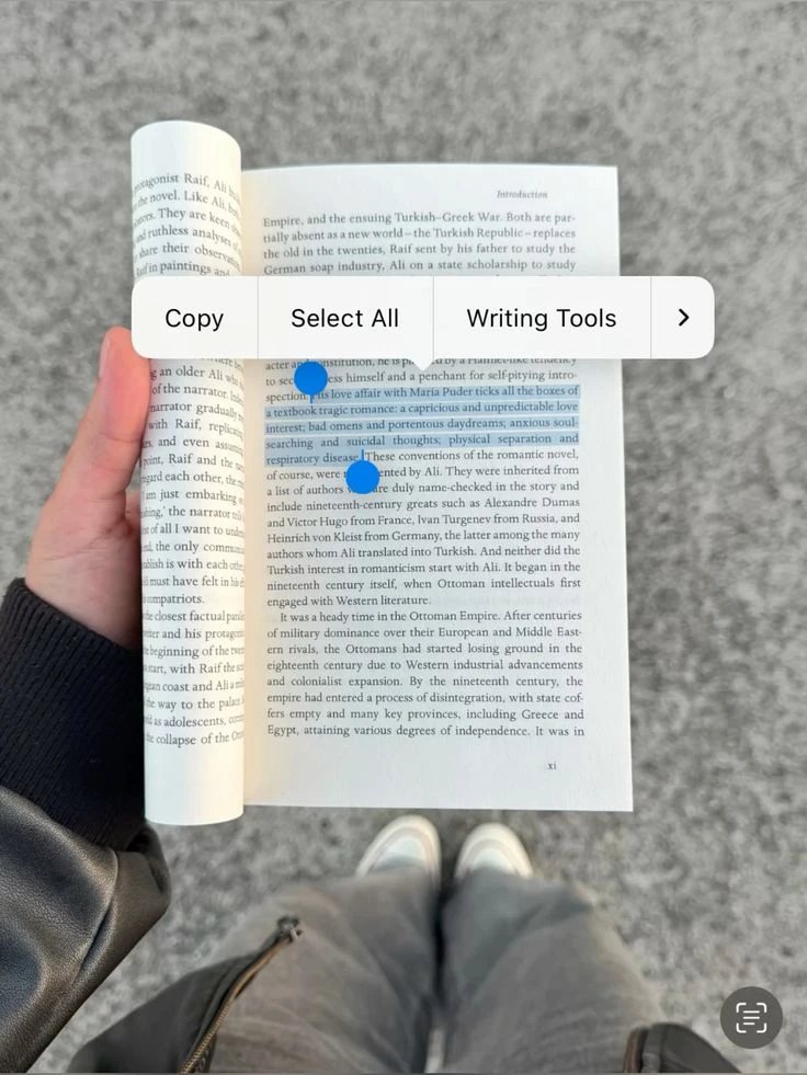 A hand holding an open paperback while a phone selects a passage of its text.