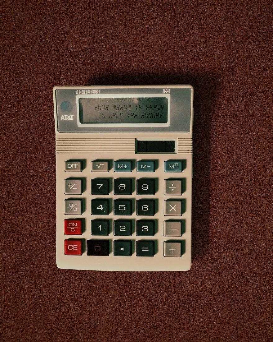 A beige desk calculator on a brown felt surface, its display reading “Your brand is ready to walk the runway.”