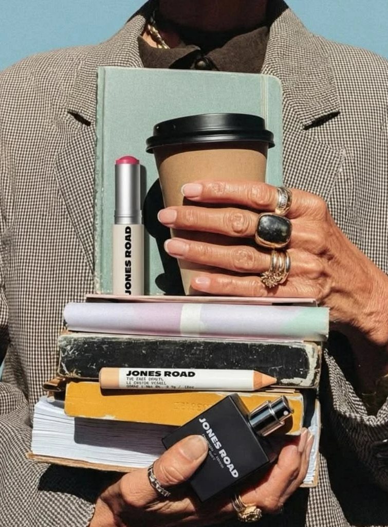 Hands with gold rings holding a stack of worn books, a coffee cup and small make-up products against a checked blazer.