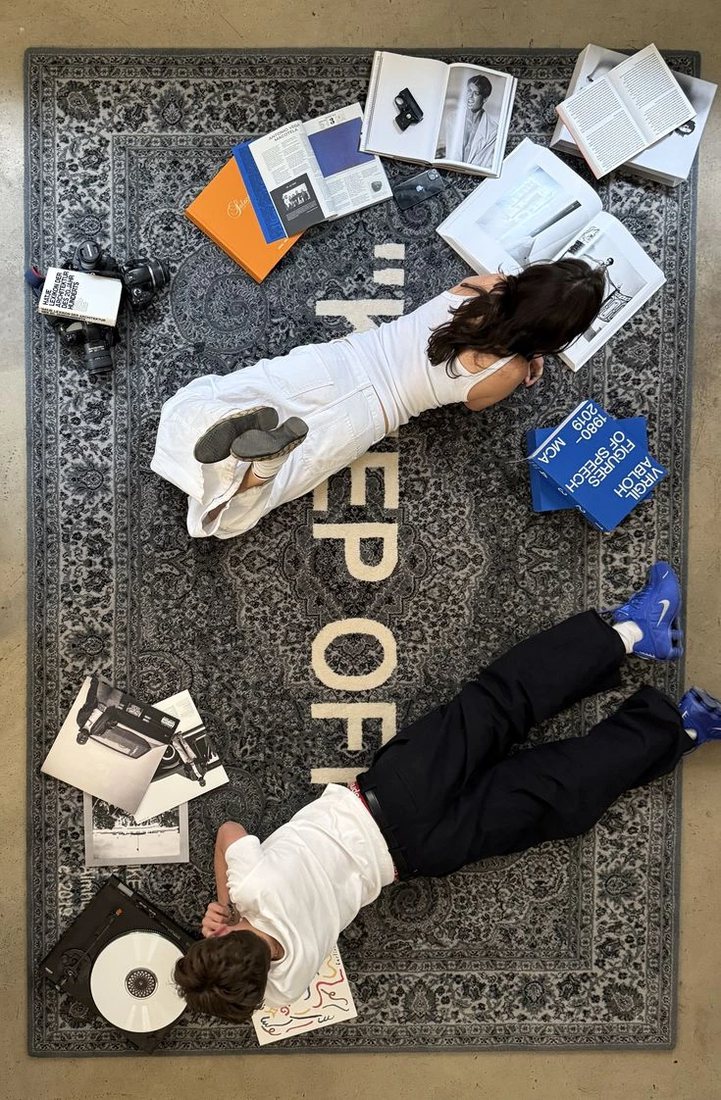 Two people lying on a patterned rug among open design books, film cameras and a record player.