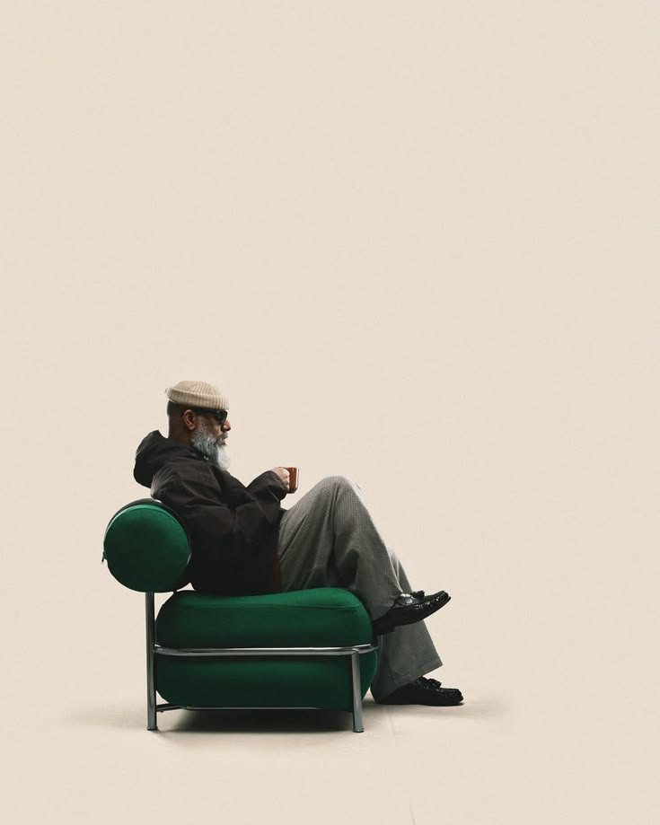 A bearded man in a beanie and sunglasses sipping coffee in a round green armchair against a cream backdrop.