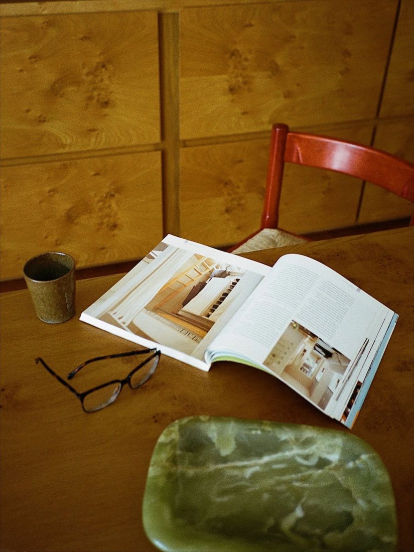 An open interiors magazine on a wooden table with glasses, a ceramic cup and a green stone dish.