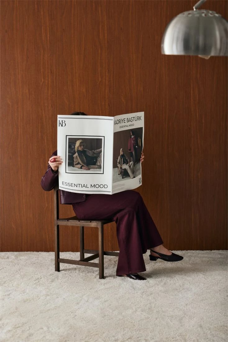 A person seated on a wooden chair against a warm wood wall, face hidden behind an open broadsheet.
