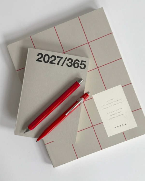 Two grey notebooks, one marked 2027/365, with two red pens on a white table.