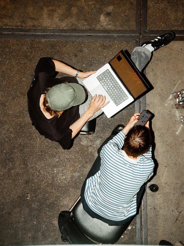 Seen from above: two people side by side on stools, one on a laptop, one on a phone.
