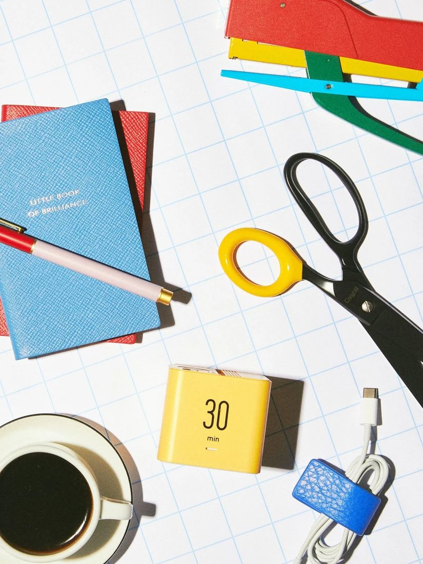 Colourful desk tools on grid paper: a blue notebook, a pen, scissors, a stapler, a yellow timer, coffee and a coiled cable.