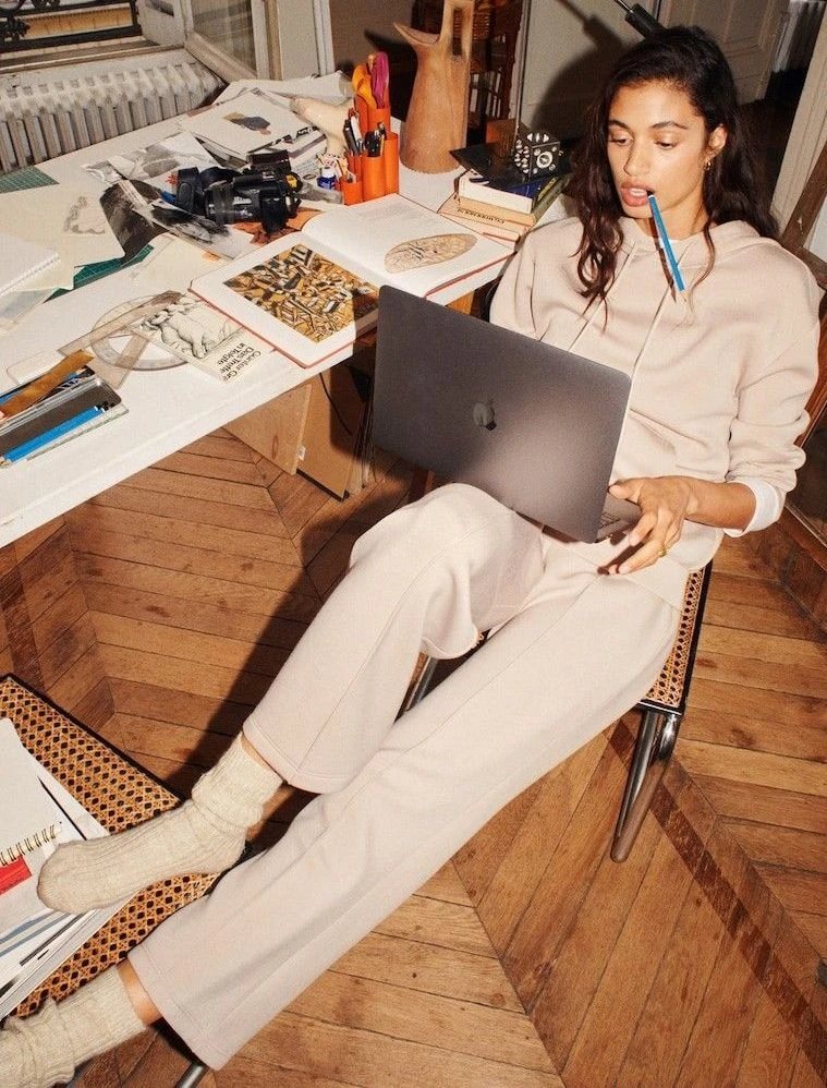 A person in a cream tracksuit leaning back from a cluttered studio desk, laptop on their lap and a pencil in their mouth.