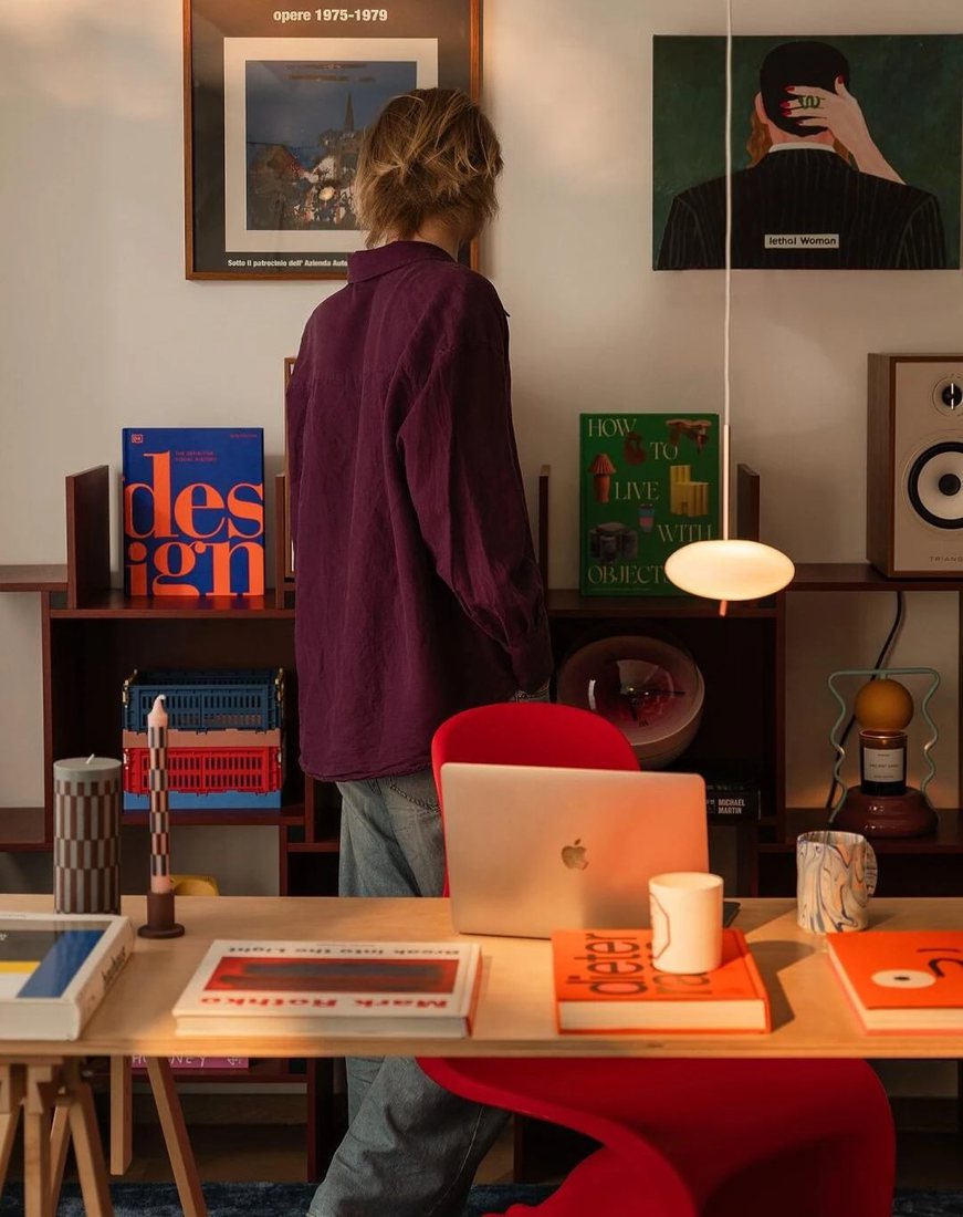 A person in a plum shirt browsing a lived-in studio: design books, a red chair, a laptop and a glowing pendant lamp.